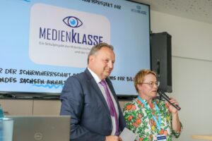 Two presenters speak at a conference in front of a large MEDIENKLASSE banner with a blue backdrop.