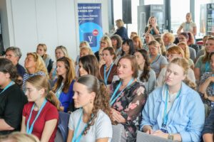 Audience of adults seated in a conference room listening to a speaker, many with blue lanyards and badges.