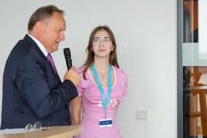 A man in a navy suit speaks into a microphone while a young woman in a pink shirt listens, wearing a lanyard with an ID badge.