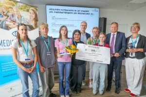 Group of students and adults posing with a large prize check at a school media-class award ceremony, with a Medienklasse banner in the background.