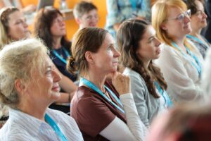 Group of adults seated in an audience at a conference, listening attentively with blue lanyards.