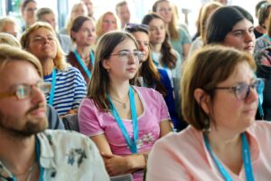 Audience of adults seated in a conference room wearing blue lanyards, listening attentively to a speaker; center foreground shows a young woman in a pink shirt and glasses.