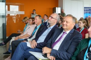 Audience of professionals seated in a conference room with an orange wall, listening attentively to a speaker, some with teal lanyards.