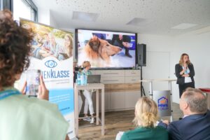 Speaker presenting at a conference in front of a large screen showing a fluffy collie; audience seated and watching.