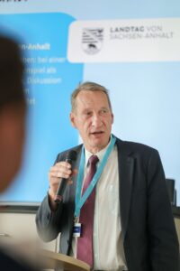 Man in a dark suit and maroon tie speaking into a handheld microphone at a conference, wearing a teal lanyard and name badge against a blue backdrop with a logo.