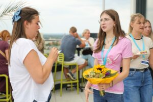Young woman in pink holding a sunflower bouquet talks with a woman in white at a rooftop social event.