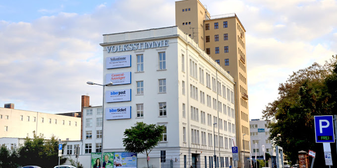 White multi-story corner building with 'VOLKESTIMME' on the façade and blue banners, next to a tall beige tower, with a street and parking signs in the foreground.